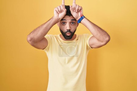 Téléchargez les photos : Homme hispanique avec barbe debout sur fond jaune faire un geste drôle avec doigt sur tête comme cornes de taureau - en image libre de droit