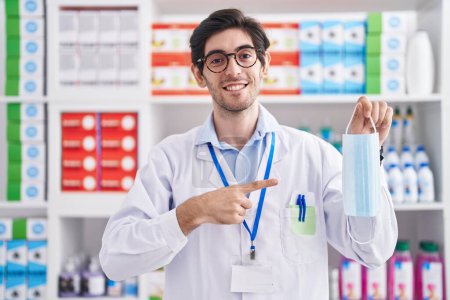 Foto de Young hispanic man working at pharmacy drugstore holding safety mask smiling happy pointing with hand and finger - Imagen libre de derechos