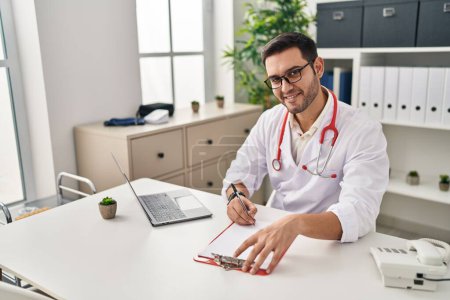 Téléchargez les photos : Young hispanic man wearing doctor uniform writing report working at clinic - en image libre de droit