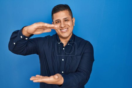 Photo for Hispanic young man standing over blue background gesturing with hands showing big and large size sign, measure symbol. smiling looking at the camera. measuring concept. - Royalty Free Image