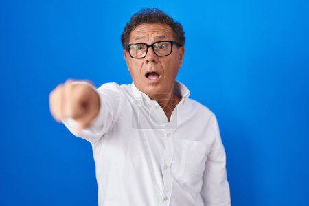 Téléchargez les photos : Middle age hispanic man standing over blue background pointing with finger surprised ahead, open mouth amazed expression, something on the front - en image libre de droit