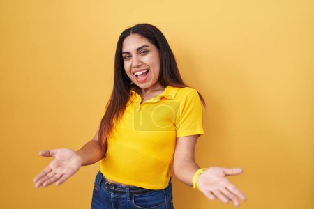 Téléchargez les photos : Jeune femme arabe debout sur fond jaune souriant gai avec les bras ouverts comme accueil amical, salutations positives et confiantes - en image libre de droit