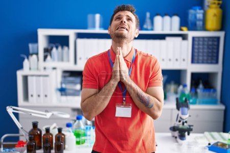 Foto de Young hispanic man working at scientist laboratory begging and praying with hands together with hope expression on face very emotional and worried. begging. - Imagen libre de derechos