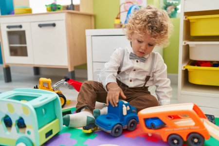 Foto de Adorable niño rubio jugando con coches juguete sentado en el suelo en el jardín de infantes - Imagen libre de derechos