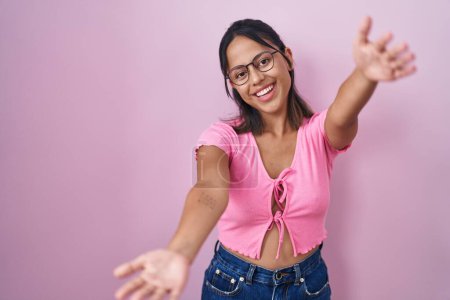 Téléchargez les photos : Jeune femme hispanique debout sur fond rose portant des lunettes regardant la caméra souriant à bras ouverts pour étreindre. expression joyeuse embrassant le bonheur. - en image libre de droit