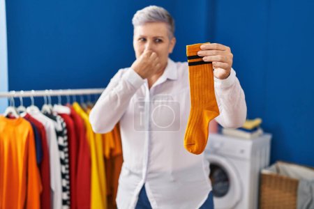 Foto de Mujer de mediana edad sonriendo confiado oliendo calcetines sucios en la sala de lavandería - Imagen libre de derechos