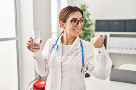 Photo for Young brunette doctor woman holding glass of water pointing thumb up to the side smiling happy with open mouth - Royalty Free Image