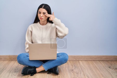 Foto de Mujer joven usando el ordenador portátil sentado en el suelo en casa disparando y matándose señalando la mano y los dedos a la cabeza como arma, gesto suicida. - Imagen libre de derechos