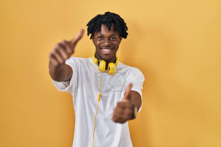 Foto de Hombre africano joven con rastas de pie sobre fondo amarillo aprobando hacer gesto positivo con la mano, pulgares arriba sonriendo y feliz por el éxito. gesto ganador. - Imagen libre de derechos