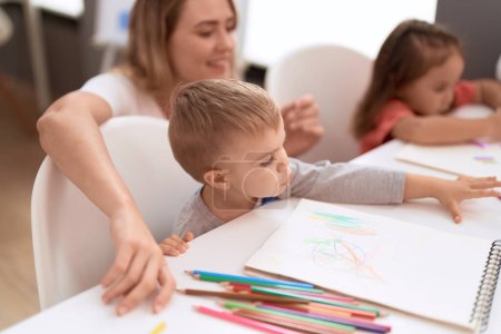 Foto de Teacher with boy and girl sitting on table drawing on paper at kindergarten - Imagen libre de derechos