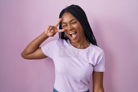 Téléchargez les photos : Femme afro-américaine avec des tresses debout sur fond rose faisant symbole de paix avec les doigts sur le visage, souriant joyeuse montrant la victoire - en image libre de droit