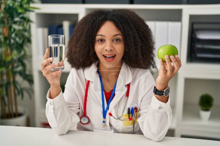Photo for Young african american dietitian woman holding fresh apple and water celebrating crazy and amazed for success with open eyes screaming excited. - Royalty Free Image