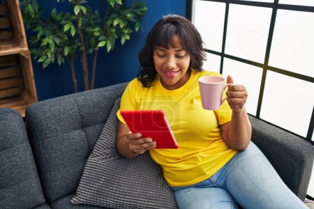 Téléchargez les photos : Jeune belle femme latine utilisant touchpad boire du café assis sur le canapé à la maison - en image libre de droit