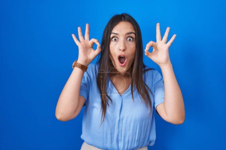 Photo for Young brunette woman standing over blue background looking surprised and shocked doing ok approval symbol with fingers. crazy expression - Royalty Free Image