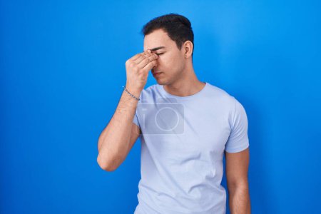 Foto de Young hispanic man standing over blue background tired rubbing nose and eyes feeling fatigue and headache. stress and frustration concept. - Imagen libre de derechos