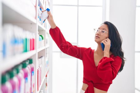 Photo for Young chinese woman customer talking on smartphone with relaxed expression at pharmacy - Royalty Free Image