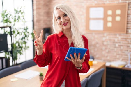 Foto de Caucasian woman working at the office with tablet smiling looking to the camera showing fingers doing victory sign. number two. - Imagen libre de derechos