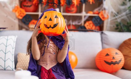 Foto de Adorable chica hispana con disfraz de halloween sosteniendo cesta de calabaza en la cara en casa - Imagen libre de derechos