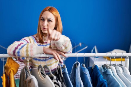 Photo for Young woman searching clothes on clothing rack pointing with finger to the camera and to you, confident gesture looking serious - Royalty Free Image