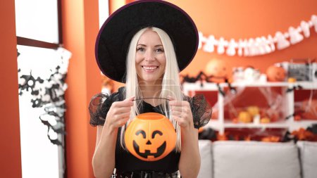 Foto de Joven mujer rubia con disfraz de bruja sosteniendo cesta de calabaza de Halloween en casa - Imagen libre de derechos