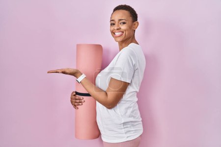 Téléchargez les photos : Belle femme afro-américaine tenant tapis de yoga pointant de côté avec les mains ouvertes paumes montrant l'espace de copie, présentant la publicité souriant heureux excité - en image libre de droit