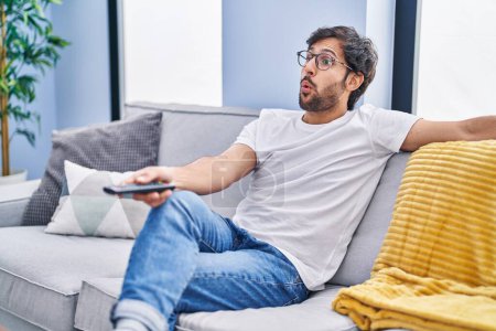 Photo for Handsome latin man holding television remote control afraid and shocked with surprise and amazed expression, fear and excited face. - Royalty Free Image