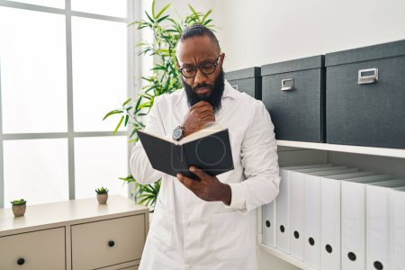 Téléchargez les photos : Young african american man wearing doctor uniform reading book at clinic - en image libre de droit