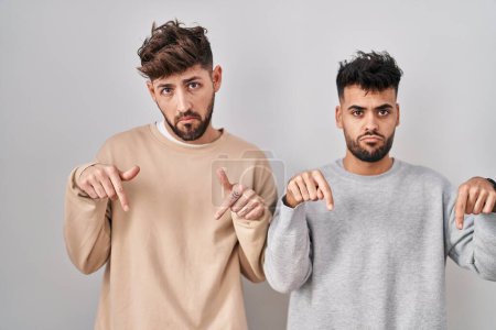 Foto de Young homosexual couple standing over white background pointing down looking sad and upset, indicating direction with fingers, unhappy and depressed. - Imagen libre de derechos