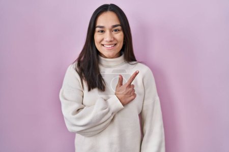 Téléchargez les photos : Young south asian woman standing over pink background cheerful with a smile on face pointing with hand and finger up to the side with happy and natural expression - en image libre de droit