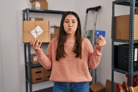 Téléchargez les photos : Young latin woman working at small business ecommerce holding credit card puffing cheeks with funny face. mouth inflated with air, catching air. - en image libre de droit