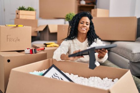 Photo for African american woman unpacking cardboard box sitting on floor at new home - Royalty Free Image