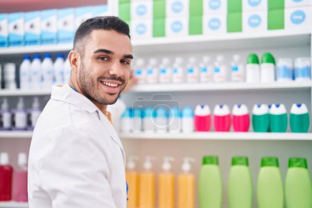 Téléchargez les photos : Jeune homme hispanique pharmacien souriant produit de maintien confiant sur les étagères à la pharmacie - en image libre de droit