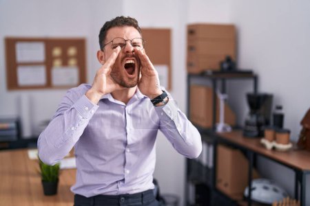 Photo for Young hispanic man at the office shouting angry out loud with hands over mouth - Royalty Free Image