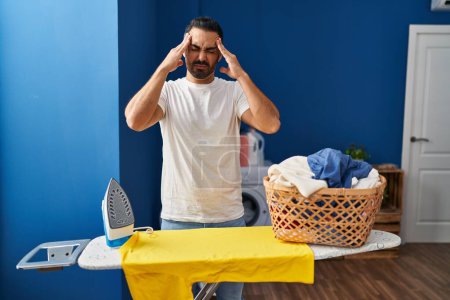 Téléchargez les photos : Young hispanic man with beard ironing clothes at home with hand on head, headache because stress. suffering migraine. - en image libre de droit