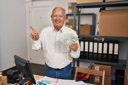 Téléchargez les photos : Homme âgé avec les cheveux gris travaillant à la petite entreprise commerce électronique tenant des dollars souriant heureux pointant avec la main et le doigt sur le côté - en image libre de droit