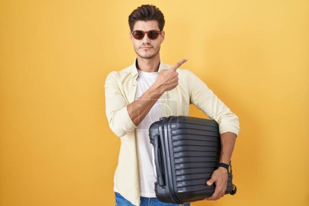 Photo for Young hispanic man holding suitcase going on summer vacation pointing with hand finger to the side showing advertisement, serious and calm face - Royalty Free Image