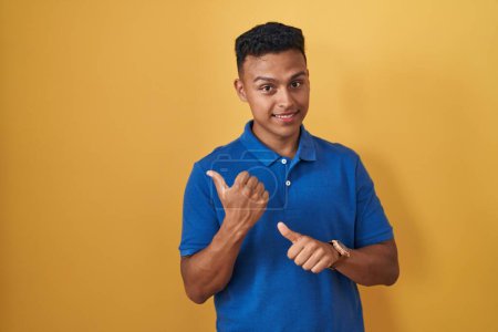Photo for Young hispanic man standing over yellow background pointing to the back behind with hand and thumbs up, smiling confident - Royalty Free Image