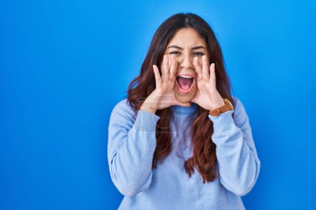 Photo for Hispanic young woman standing over blue background shouting angry out loud with hands over mouth - Royalty Free Image