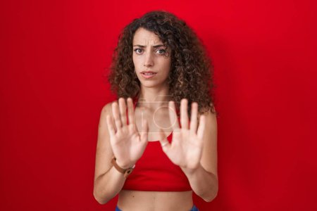 Téléchargez les photos : Hispanic woman with curly hair standing over red background moving away hands palms showing refusal and denial with afraid and disgusting expression. stop and forbidden. - en image libre de droit