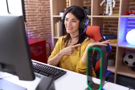 Foto de Middle age hispanic woman playing video games using headphones amazed and smiling to the camera while presenting with hand and pointing with finger. - Imagen libre de derechos