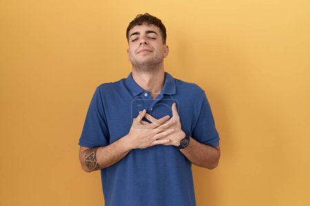 Photo for Young hispanic man standing over yellow background smiling with hands on chest with closed eyes and grateful gesture on face. health concept. - Royalty Free Image