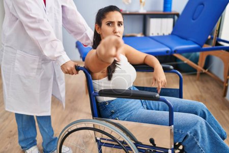 Téléchargez les photos : Young hispanic woman sitting on wheelchair at physiotherapy clinic pointing with finger to the camera and to you, confident gesture looking serious - en image libre de droit