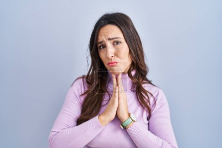 Photo for Young brunette woman standing over blue background begging and praying with hands together with hope expression on face very emotional and worried. begging. - Royalty Free Image