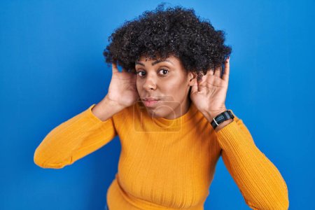 Photo for Black woman with curly hair standing over blue background trying to hear both hands on ear gesture, curious for gossip. hearing problem, deaf - Royalty Free Image