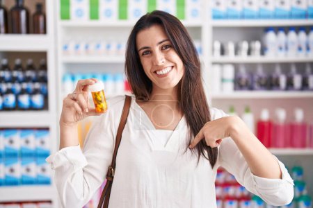 Foto de Young brunette woman shopping at pharmacy drugstore holding pills pointing finger to one self smiling happy and proud - Imagen libre de derechos