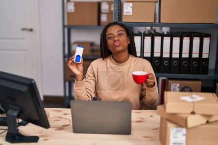 Foto de Young african american with braids working at small business ecommerce drinking a coffee with sweetener looking at the camera blowing a kiss being lovely and sexy. love expression. - Imagen libre de derechos
