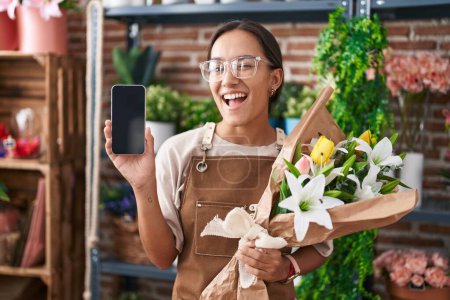 Foto de Mujer hispana joven que trabaja en floristería mostrando la pantalla del teléfono inteligente guiñando el ojo a la cámara con expresión sexy, cara alegre y feliz. - Imagen libre de derechos