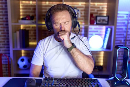 Téléchargez les photos : Homme d'âge moyen avec barbe jouant à des jeux vidéo portant des écouteurs se sentant mal et toussant comme symptôme de rhume ou de bronchite. concept de soins de santé. - en image libre de droit