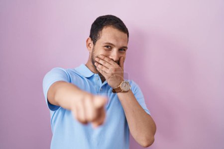 Téléchargez les photos : Hispanic man standing over pink background laughing at you, pointing finger to the camera with hand over mouth, shame expression - en image libre de droit