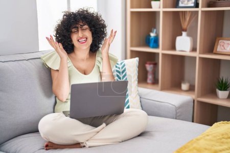 Téléchargez les photos : Jeune femme brune aux cheveux bouclés utilisant un ordinateur portable assis sur le canapé à la maison célébrant fou et fou de succès avec les bras levés et les yeux fermés hurlant excités. concept gagnant - en image libre de droit